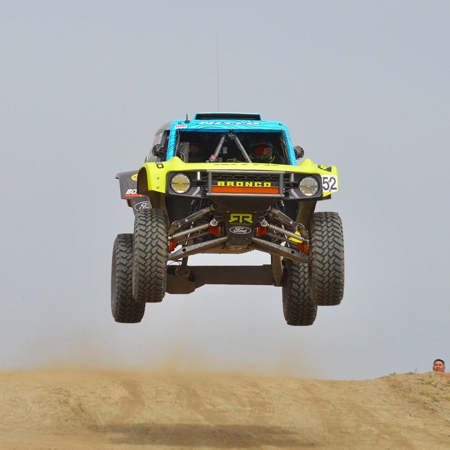 360-Series Round Lights by Rigid: A blue and yellow Ford Bronco race truck with round lights in its front bumper jumps high over a dirt crest.