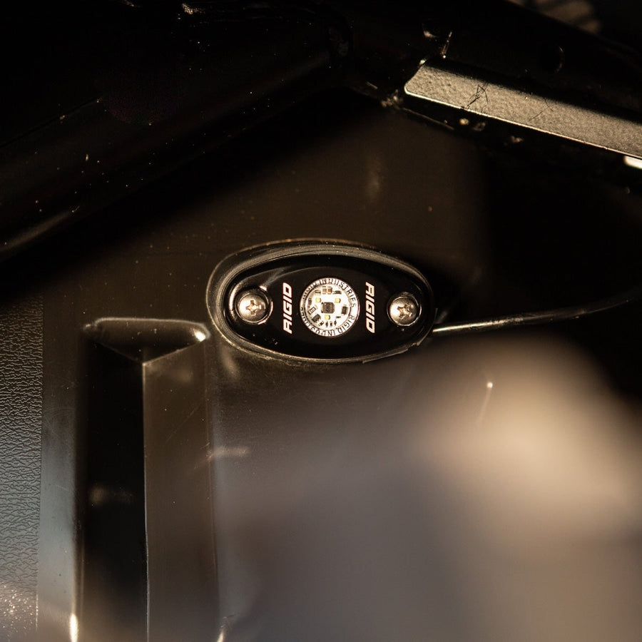 A-Series Rock & Utility Lights by Rigid: A black oval rock light mounted on the underside of a metal panel casts a soft white glow.