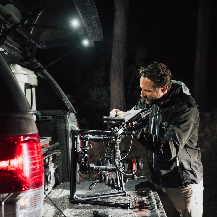 A-Series Rock & Utility Lights by Rigid: At night a man adjusts a camera gimbal at the open rear of an SUV lit by small white lights under the liftgate.