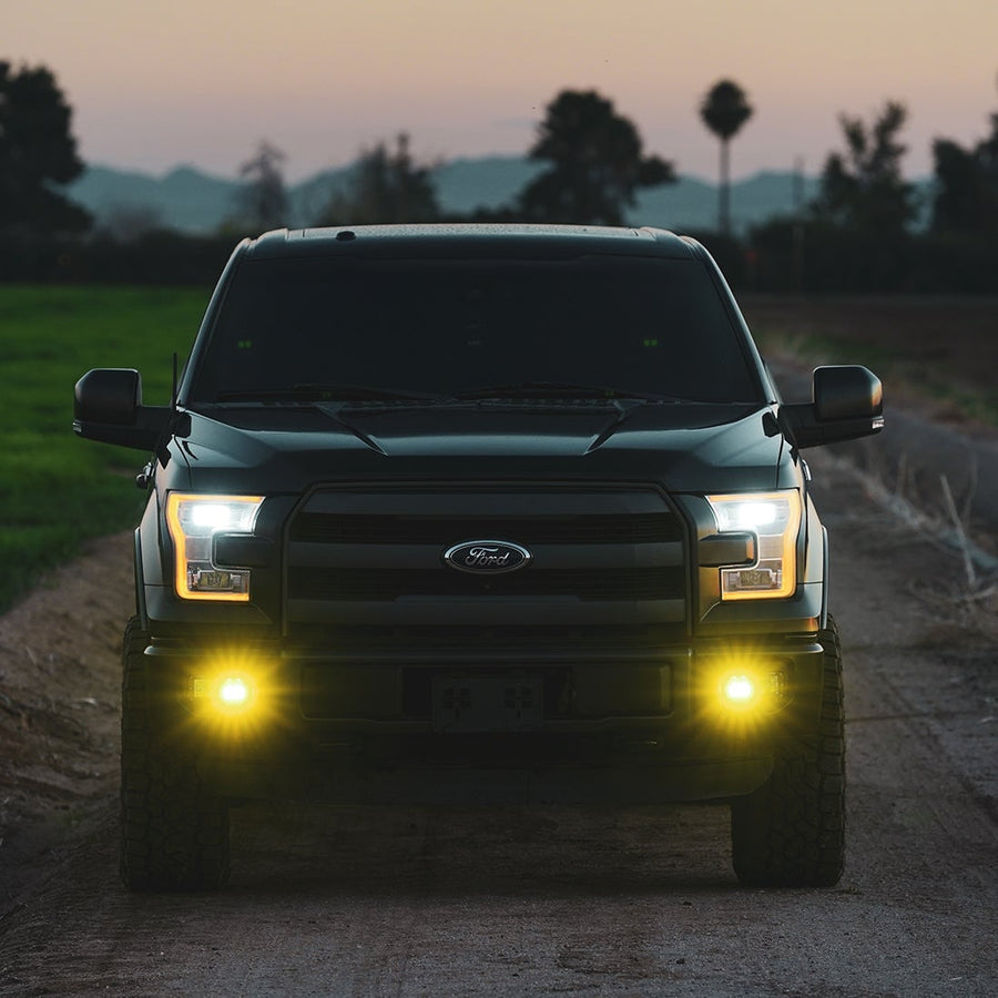 D-Series Pods by Rigid: A dark Ford F-150 parked on a dirt road at dusk with yellow SAE fog pods lit in its fog pockets.