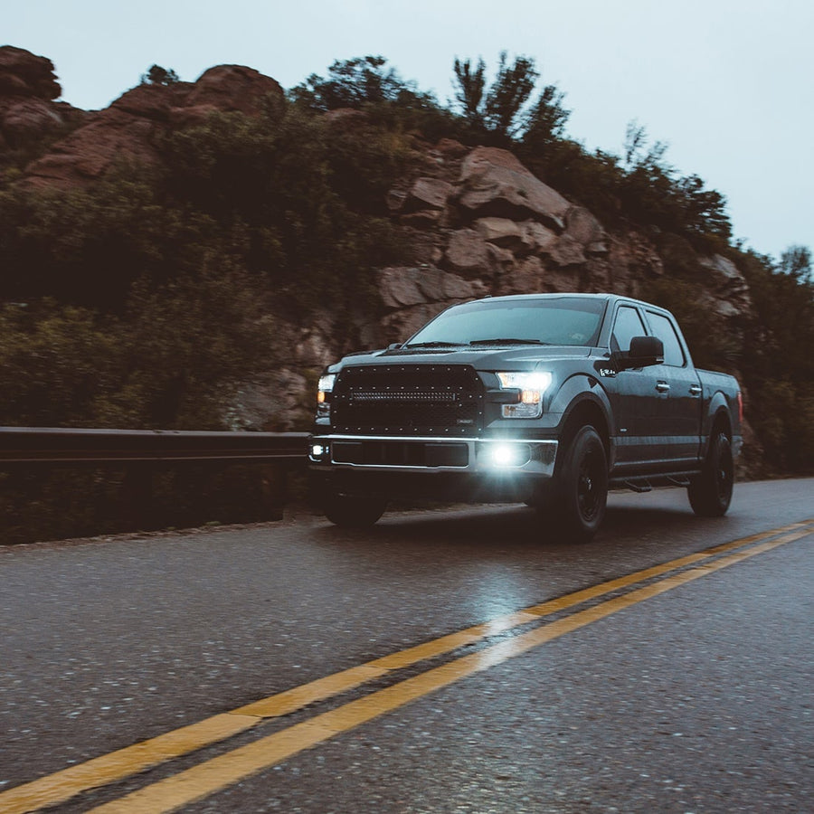 D-Series Pods by Rigid: A dark Ford F-150 drives a winding canyon road with white fog pods lit in its lower bumper.
