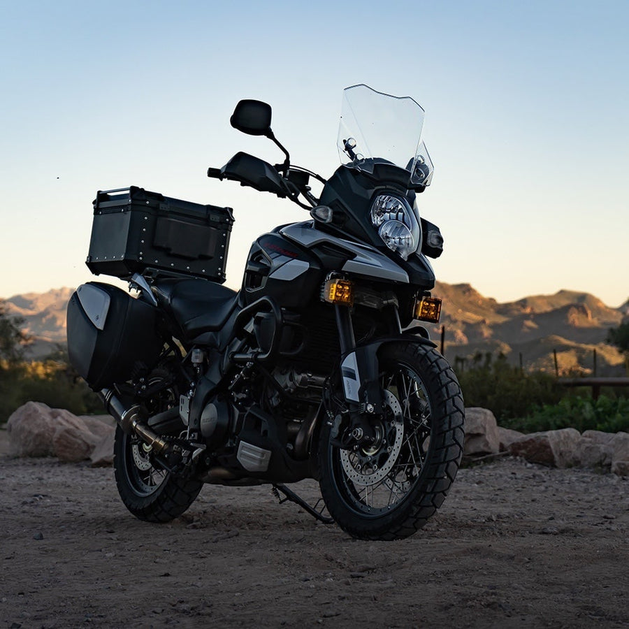D-Series Pods by Rigid: A black adventure motorcycle with luggage and small amber auxiliary lights parked in the desert at sunset.