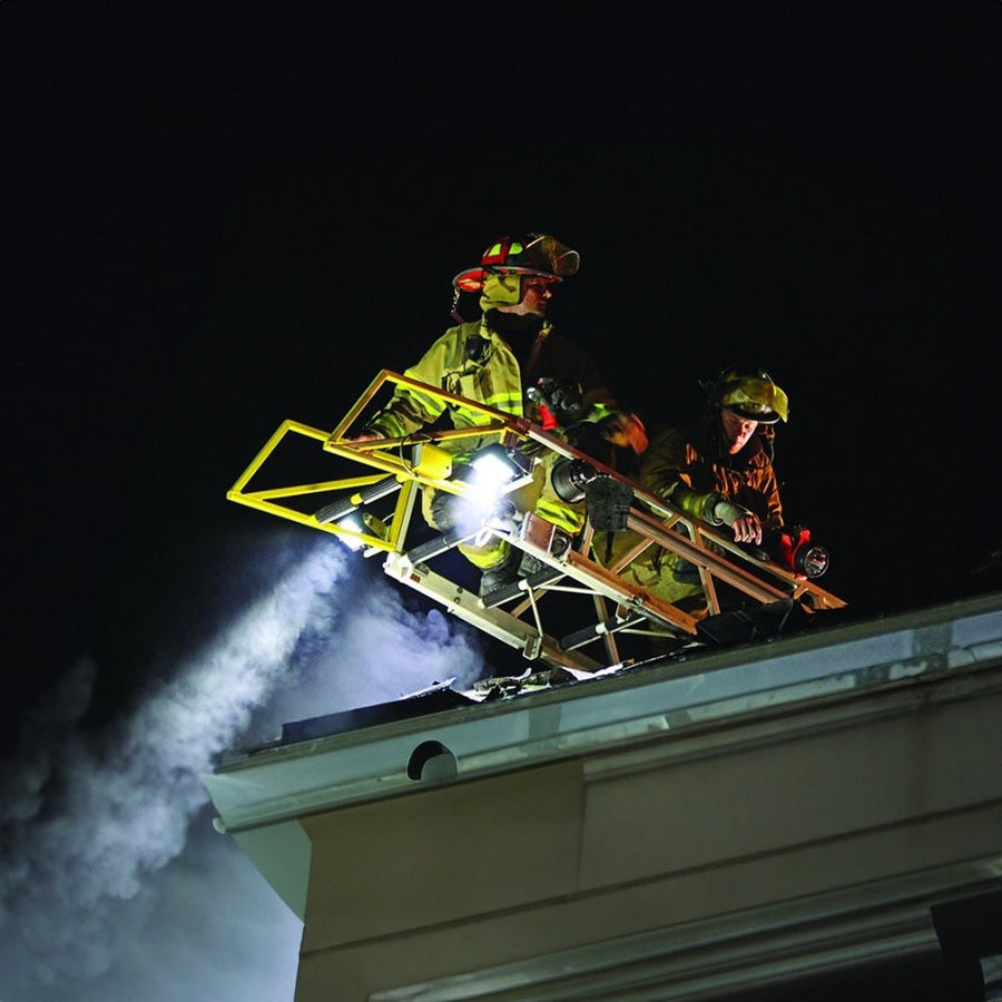 Q-Series Lights by Rigid: Two firefighters on an aerial ladder at a roofline use a bright work light to cut through smoke at night.