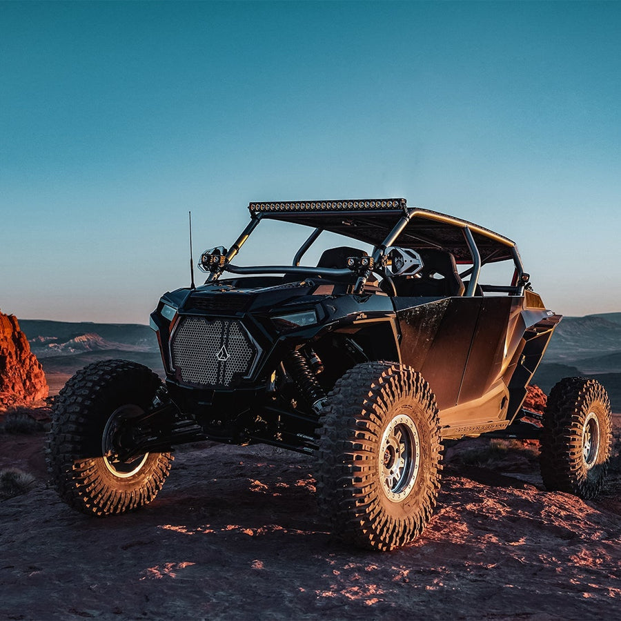 Revolve Lights by Rigid: A black Polaris RZR four-seat side-by-side on rocky ground at dusk with a roof-mounted light bar.