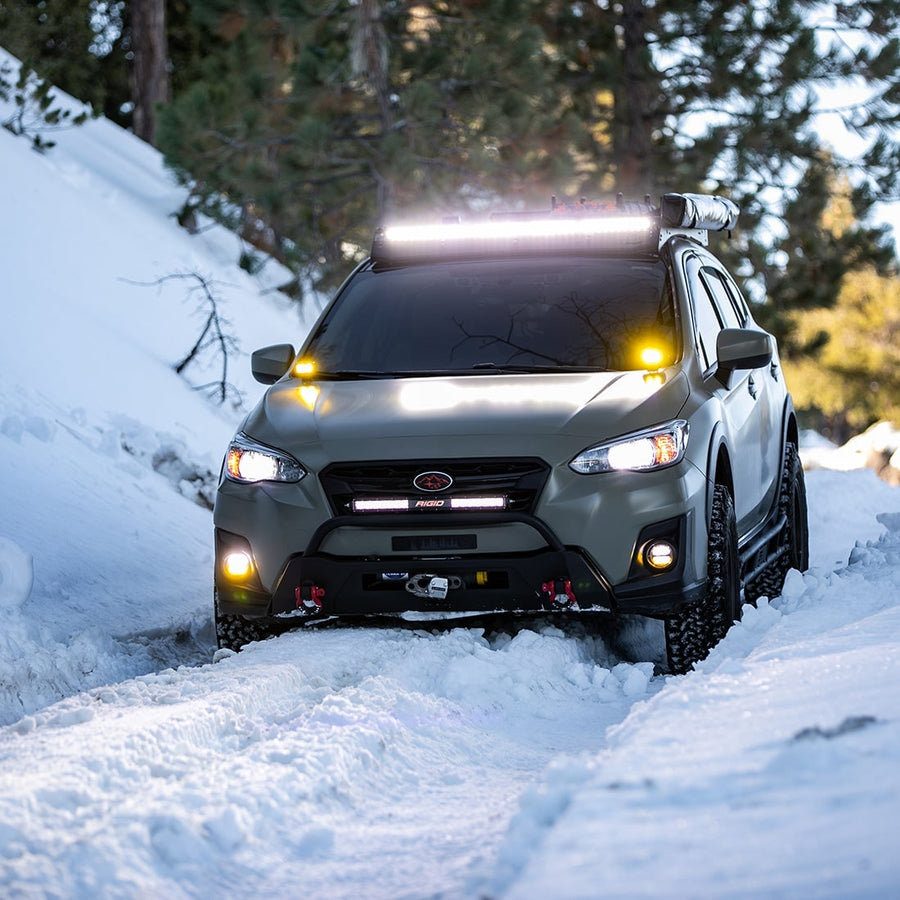 SR-Series Light Bars by Rigid: A green Subaru Crosstrek climbs a snowy trail with a lit roof light bar and amber fog lights.