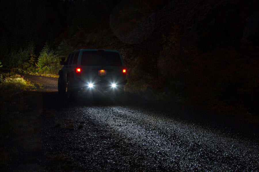 Light Mounts & Hardware by Rigid: An SUV on a dark forest trail lights the ground behind it with bright rear lights.