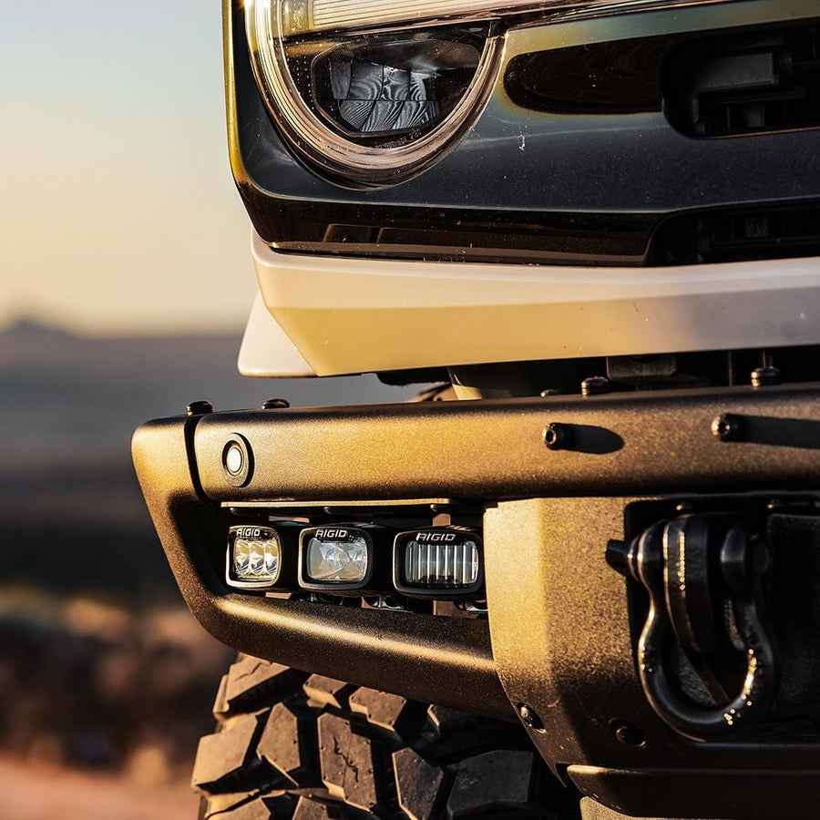 SR-M & SR-Q Pods by Rigid: Three Rigid SR-M pods sit in the light pocket of a textured black steel bumper below a round headlight at sunset.