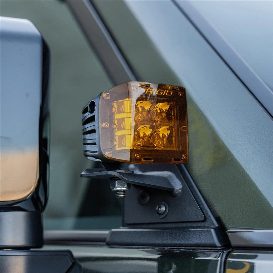 Vehicle-Specific Light Kits by Rigid: D-SS pod wearing an amber lens cover mounted at the A-pillar of a dark green vehicle; amber cover may not be included in the spot kit