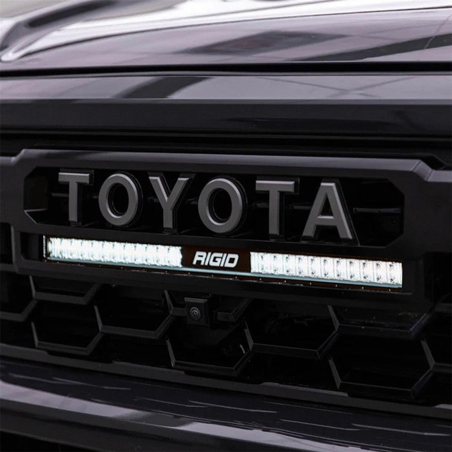 Vehicle-Specific Light Kits by Rigid: Close-up of a lit 20 inch SR-Series light bar installed under the TOYOTA lettering in a black Tacoma grille
