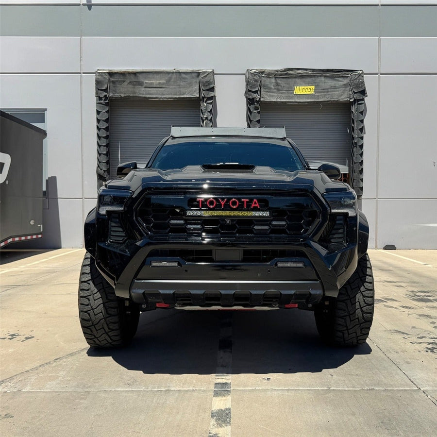 Vehicle-Specific Light Kits by Rigid: Front view of a black lifted Toyota Tacoma with red TOYOTA grille lettering and a 20 inch light bar in the grille, parked by loading docks