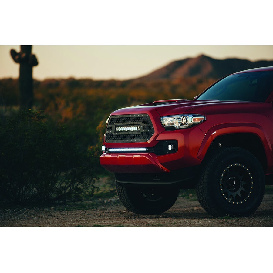 SR-Series Light Bars by Rigid: A red Toyota Tacoma with a lit Rigid light bar in the lower bumper and grille bar sits in the desert at dusk.