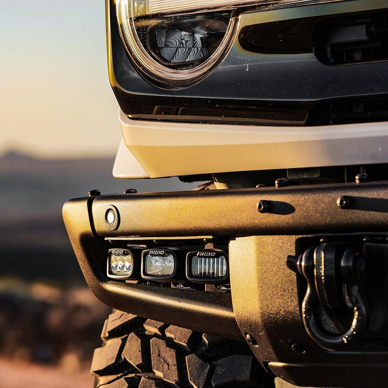 Rigid SR-M Series PRO Spot Pod installed on a Ford Bronco: three Rigid SR-M pods sit in the light pocket of a textured black steel bumper below a round headlight at sunset