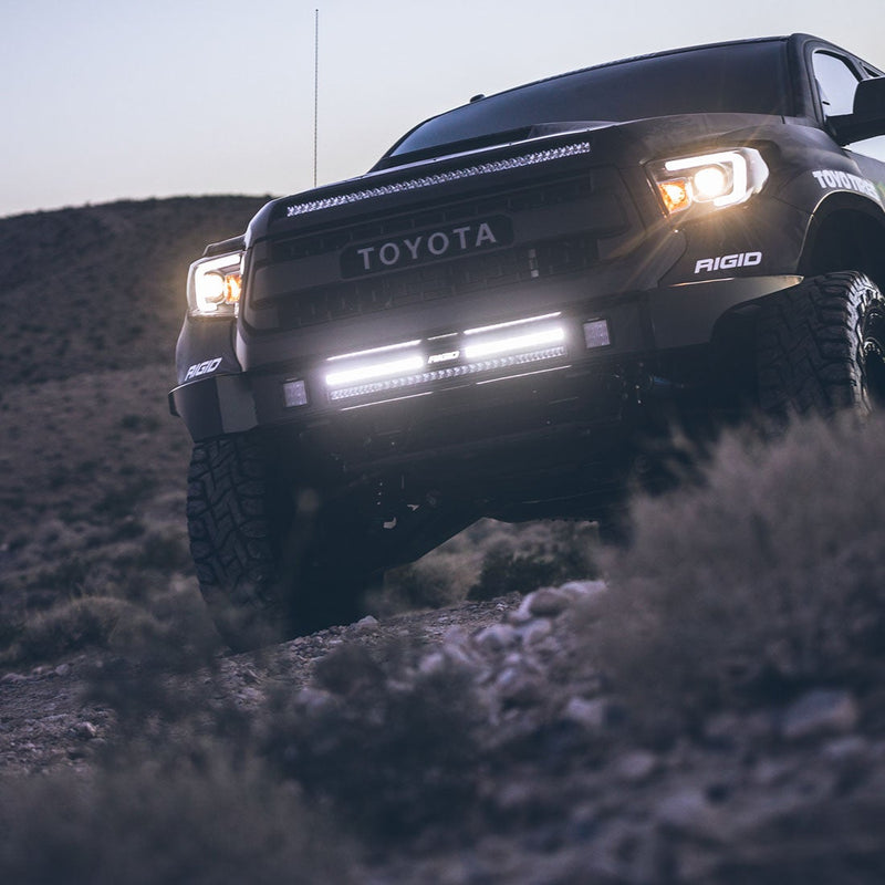 Rigid SR-Series 30" Dual Function SAE with White Backlight installed on a Toyota Tundra: A black Toyota Tundra crawls over rocks at dusk with a lit light bar mounted in its front bumper