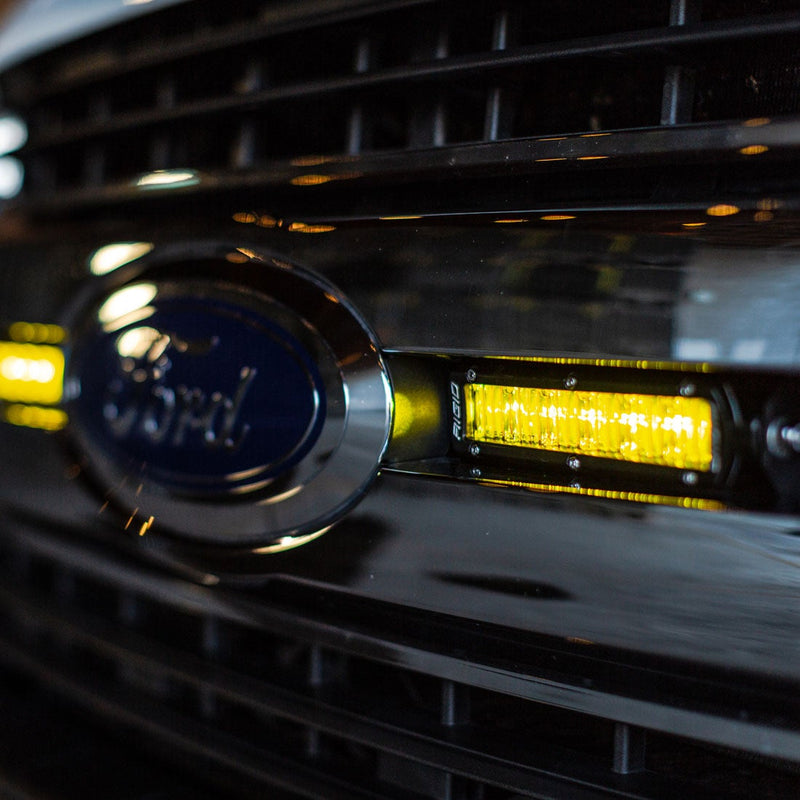 Rigid SR-Series 6" SAE Fog Lights Selective Yellow Pair installed on a Ford Super Duty: close-up of a lit selective yellow 6" SR-Series light mounted in a chrome Ford grille next to the Ford oval badge