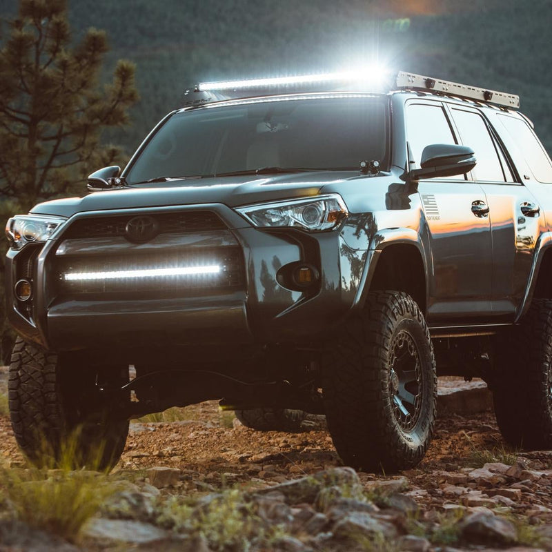 Rigid SR-Series PRO 20" Spot/Flood Combo installed on a Toyota 4Runner: A gray Toyota 4Runner on a rocky trail shows a lit Rigid bar in the lower grille and another on the roof rack