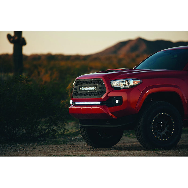 Rigid SR-Series PRO 6" Spot installed on a Toyota Tacoma: A red Toyota Tacoma with a lit Rigid light bar in the lower bumper and grille bar sits in the desert at dusk