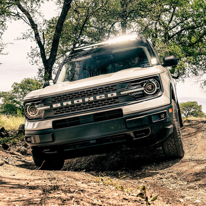 Rigid 360-Series 4" Drive A-Pillar Light Kit: white Ford Bronco Sport with BRONCO grille lettering climbing a dirt trail with lit A-pillar pods and a lit roof bar, a male driver behind the windshield