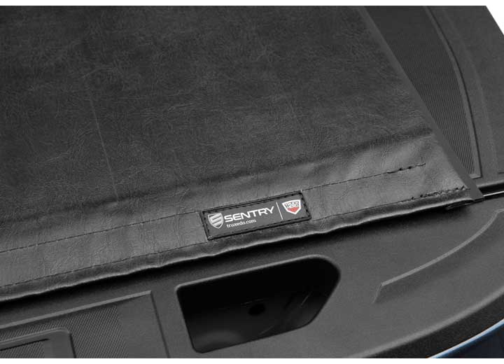 Close-up of the SENTRY name plate at the cover's rear corner, seen obliquely across the handle recess on a TruXedo Sentry hard roll-up tonneau cover