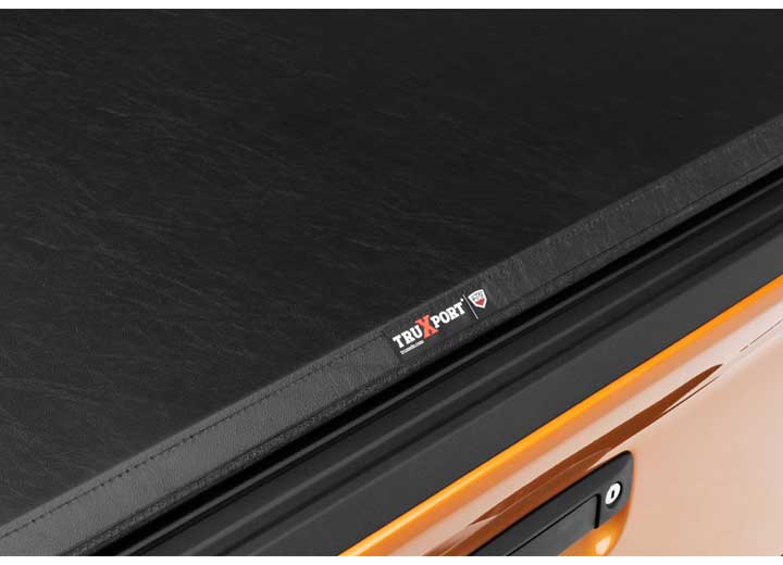 Close-up of the TRUXPORT name plate small along the cover's rear edge, above an orange bedside on a TruXedo TruXport soft roll-up tonneau cover