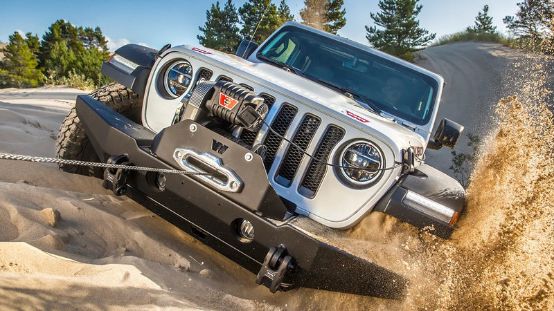 White Jeep Wrangler winching through sand with a Warn M8274-S winch in the front bumper, synthetic rope running through a polished hawse fairlead