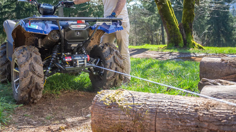 A Warn R-Series series steel-rope winch in use on a utility machine, wire rope running over a roller fairlead to a log