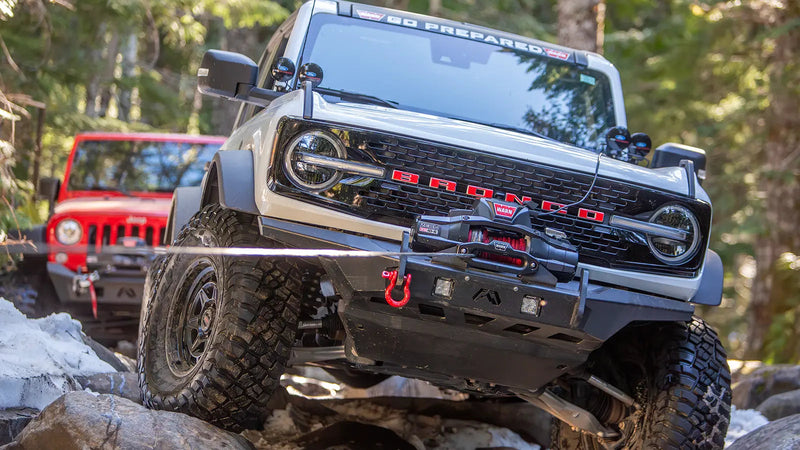 Warn ZEON XD winch pulling a Ford Bronco up a rock ledge, with a red Jeep Wrangler waiting behind
