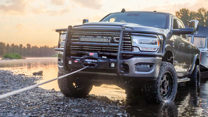 Warn ZEON XD winch pulling line from the front bumper of a Ram heavy-duty pickup on a river bank at sunrise