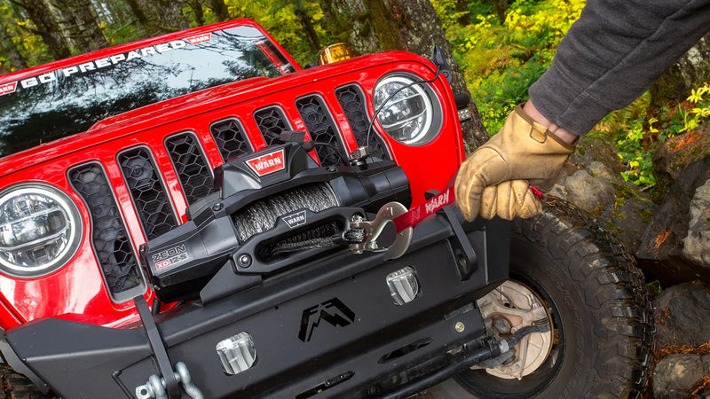 Gloved hand pulling Spydura synthetic rope from a Warn ZEON XD winch mounted in the bumper of a red Jeep Wrangler