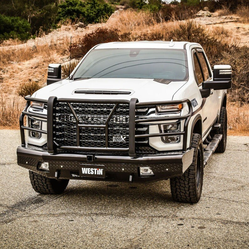Three quarter angle view of the black Westin HDX Bandit front bumper and grille guard installed on a white Chevrolet Silverado HD parked on gravel