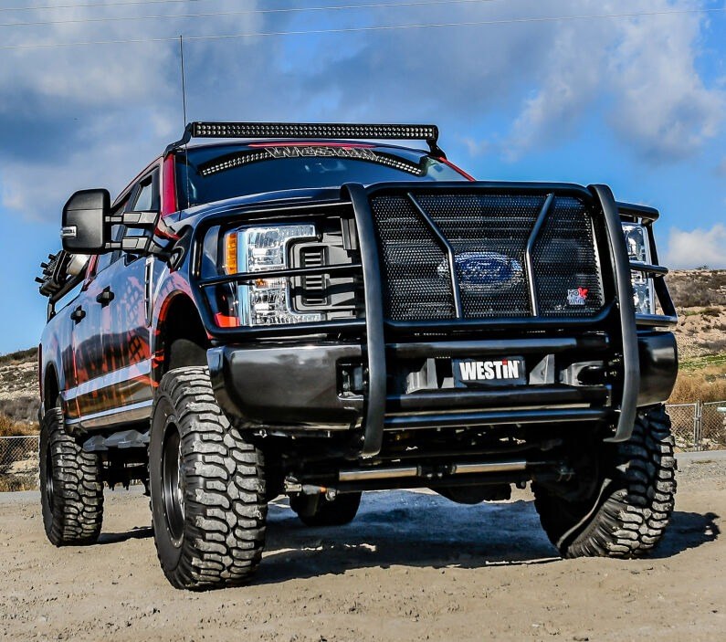 Black textured Westin HDX grille guard installed on a lifted black Ford Super Duty pickup parked on dirt under a blue sky