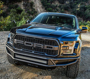 Front-on elevated view of the Westin Outlaw front off-road bumper in black with a mounted LED light bar and Westin badge on a black Ford F-150 Raptor on a dirt trail