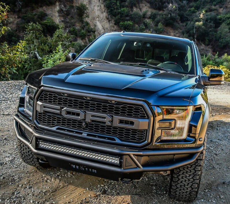Front-on elevated view of the Westin Outlaw front off-road bumper in black with a mounted LED light bar and Westin badge on a black Ford F-150 Raptor on a dirt trail