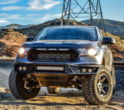 Westin Outlaw front off-road bumper in black with a centre LED light bar, four round auxiliary lights and a skid plate fitted to a silver Ford Ranger on a dirt road beside railway tracks