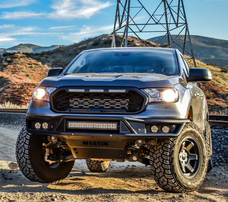 Westin Outlaw front off-road bumper in black with a centre LED light bar, four round auxiliary lights and a skid plate fitted to a silver Ford Ranger on a dirt road beside railway tracks