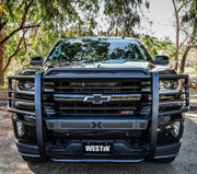 Head-on front view of a black Chevrolet Silverado fitted with a Westin Sportsman X grille guard in black, showing the wrap-around uprights, mesh punch plate and Westin-branded license plate cover