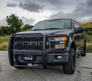 Westin Sportsman X grille guard in black installed on a black Ford F-150 with amber grille lights, photographed from a front three-quarter angle on a gravel lot under a cloudy sky