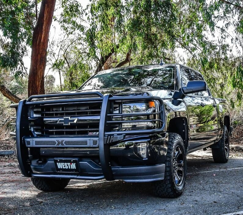 Westin Sportsman X grille guard in textured black mounted on the front of a black Chevrolet Silverado pickup parked under eucalyptus trees, shown from a front three-quarter angle