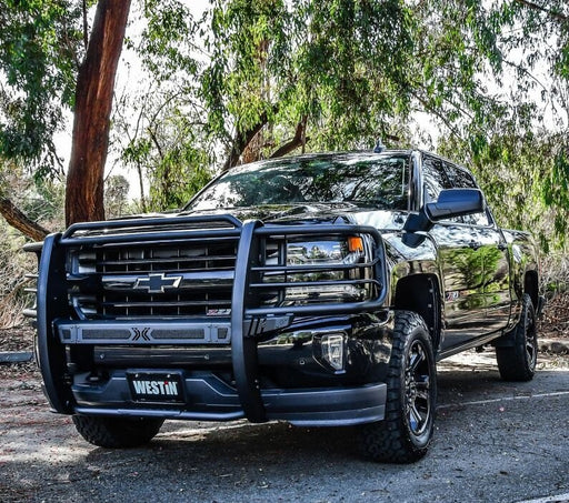 Westin Sportsman X grille guard in textured black mounted on the front of a black Chevrolet Silverado pickup parked under eucalyptus trees, shown from a front three-quarter angle