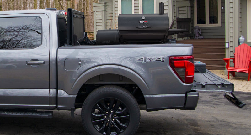 Worksport NEXUS hard folding tonneau cover shown on a 2024+ Ford F-150, a newer generation than the trucks this listing fits: cover flipped up with a smoker grill in the bed of a gray F-150