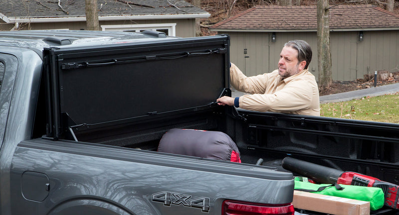 Worksport NEXUS hard folding tonneau cover on a pickup: a man reaches into the bed of a gray Ford F-150 4x4 with the cover flipped up and gear loaded inside