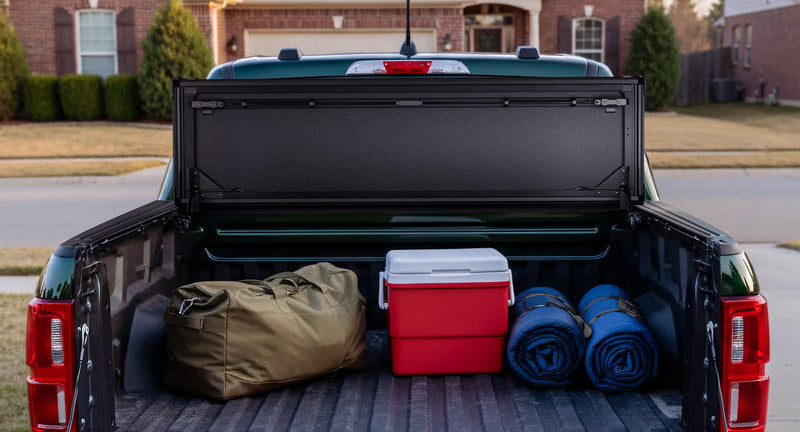 Worksport NEXUS hard folding tonneau cover on a pickup: rear view into a pickup bed with the cover flipped up behind the cab and a duffel, red cooler and sleeping pads on the floor, houses behind