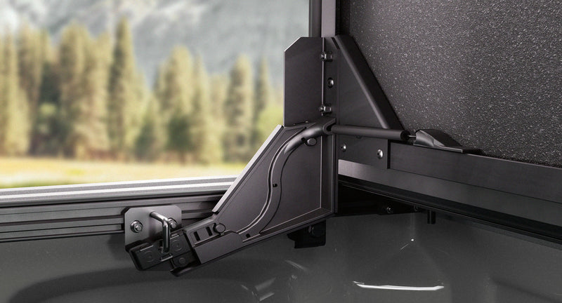 Close-up of the Worksport NEXUS hard folding tonneau cover: close-up of the black auto-locking support arm holding the flipped-up front panel upright on the bed rail, trees in the background