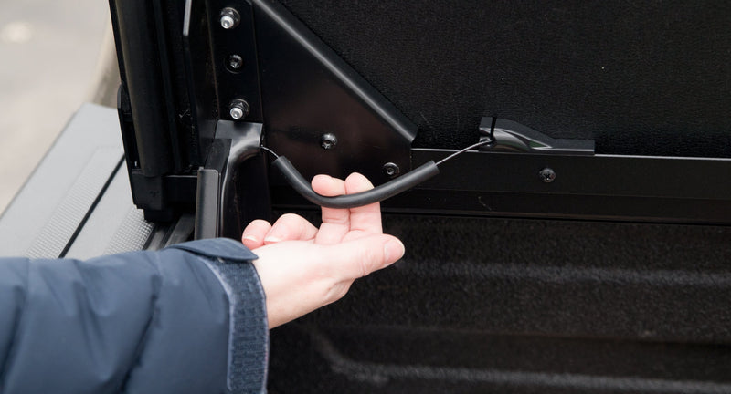 Close-up of the Worksport NEXUS hard folding tonneau cover: close-up of a hand pulling the release cable beside the auto-locking arm under the flipped-up front panel